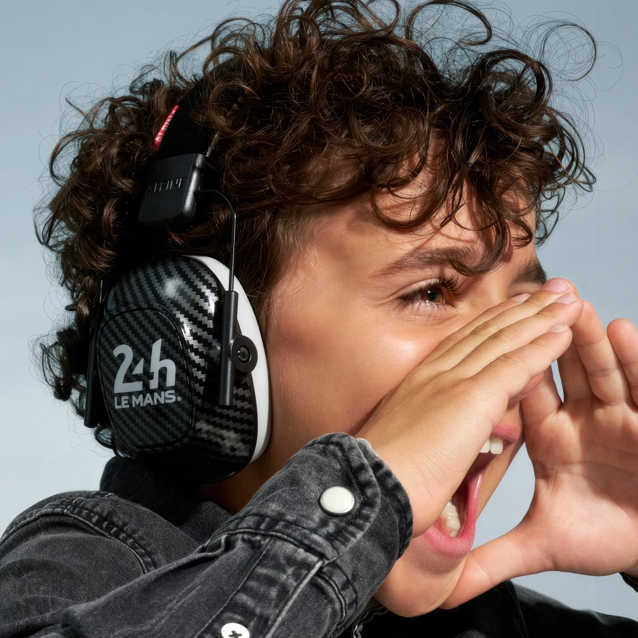 Child with curly hair cheering with hands around mouth, wearing Alpine 24h Le Mans Muffy earmuffs 